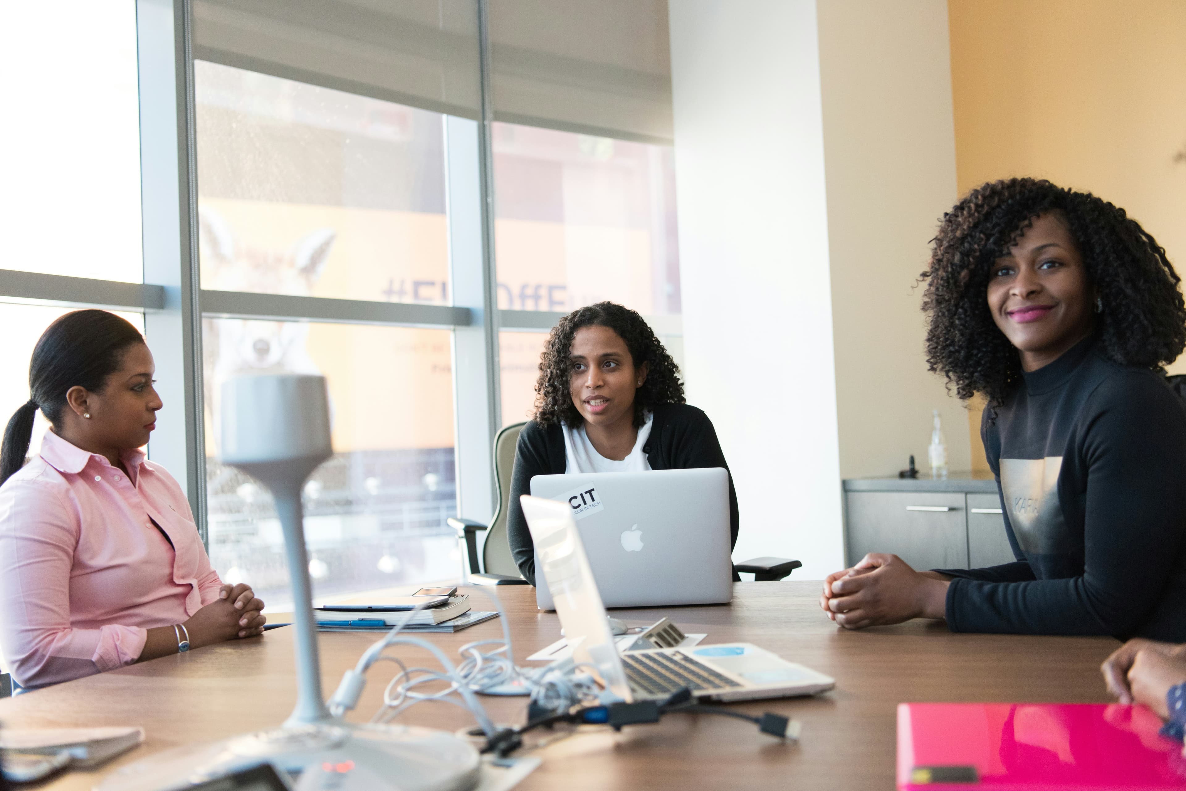 Three women meeting around a conference table with laptops in a bright office.
