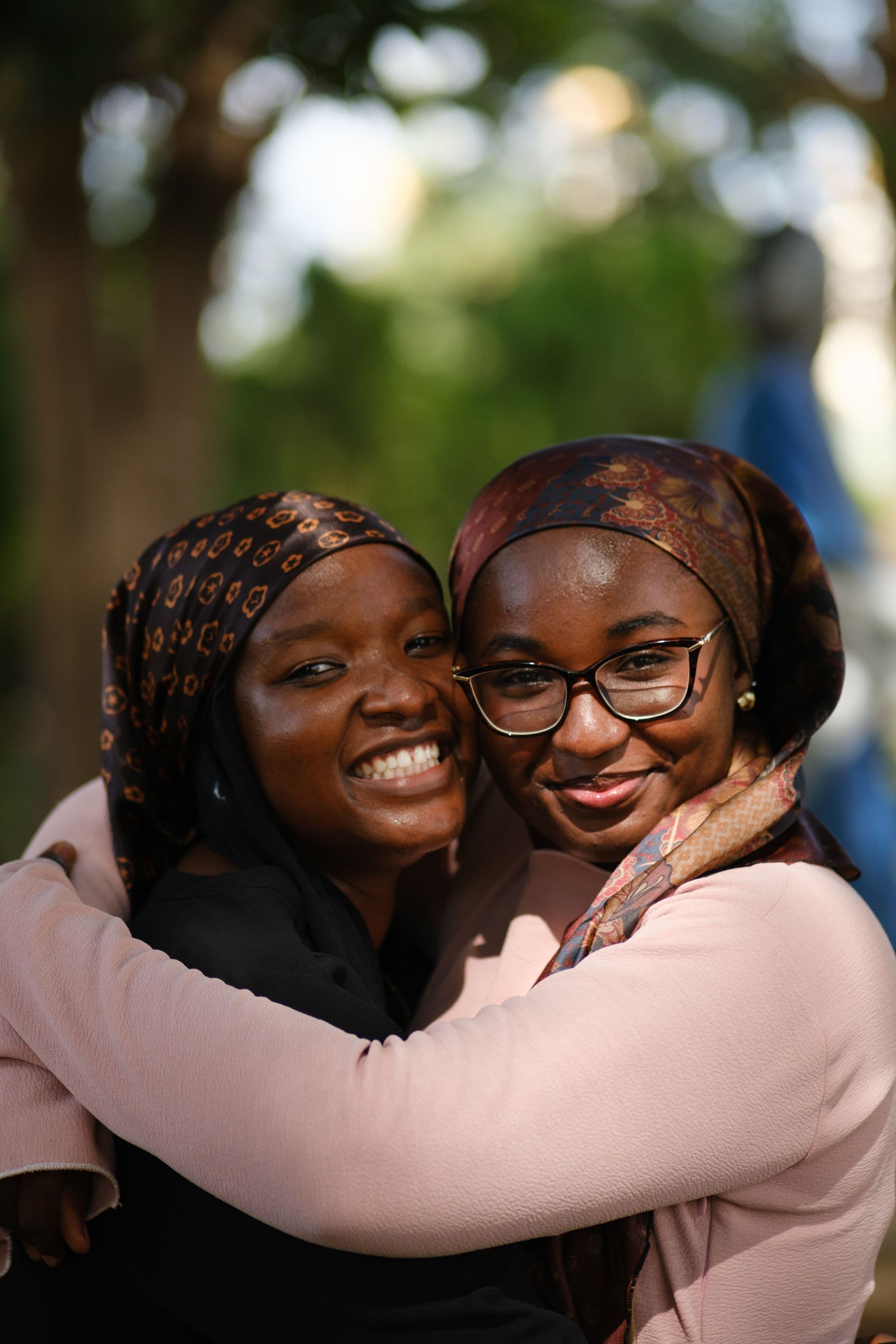 Two women smiling with their arms around each other in a sunlit park.