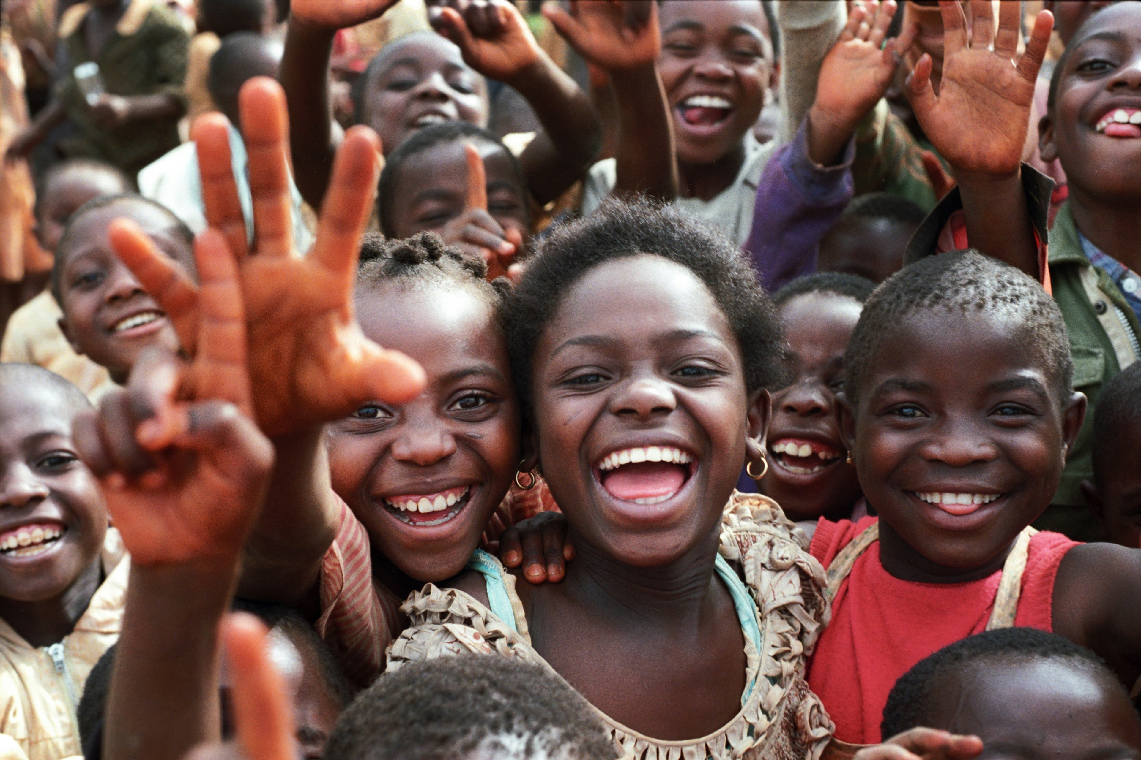 A crowd of children smiling and waving, gathered closely together.