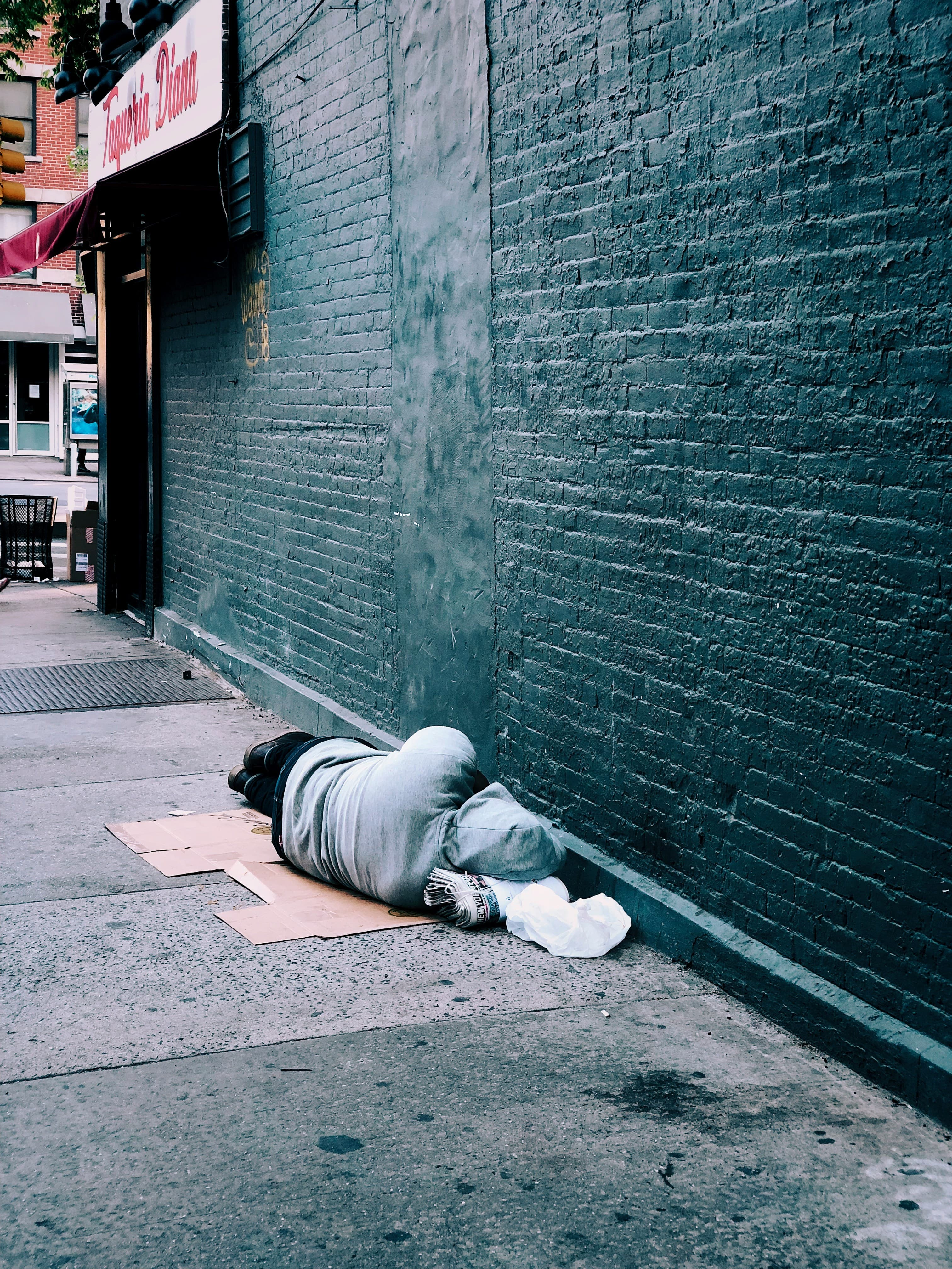 A person lying on cardboard on a sidewalk beside a brick wall.