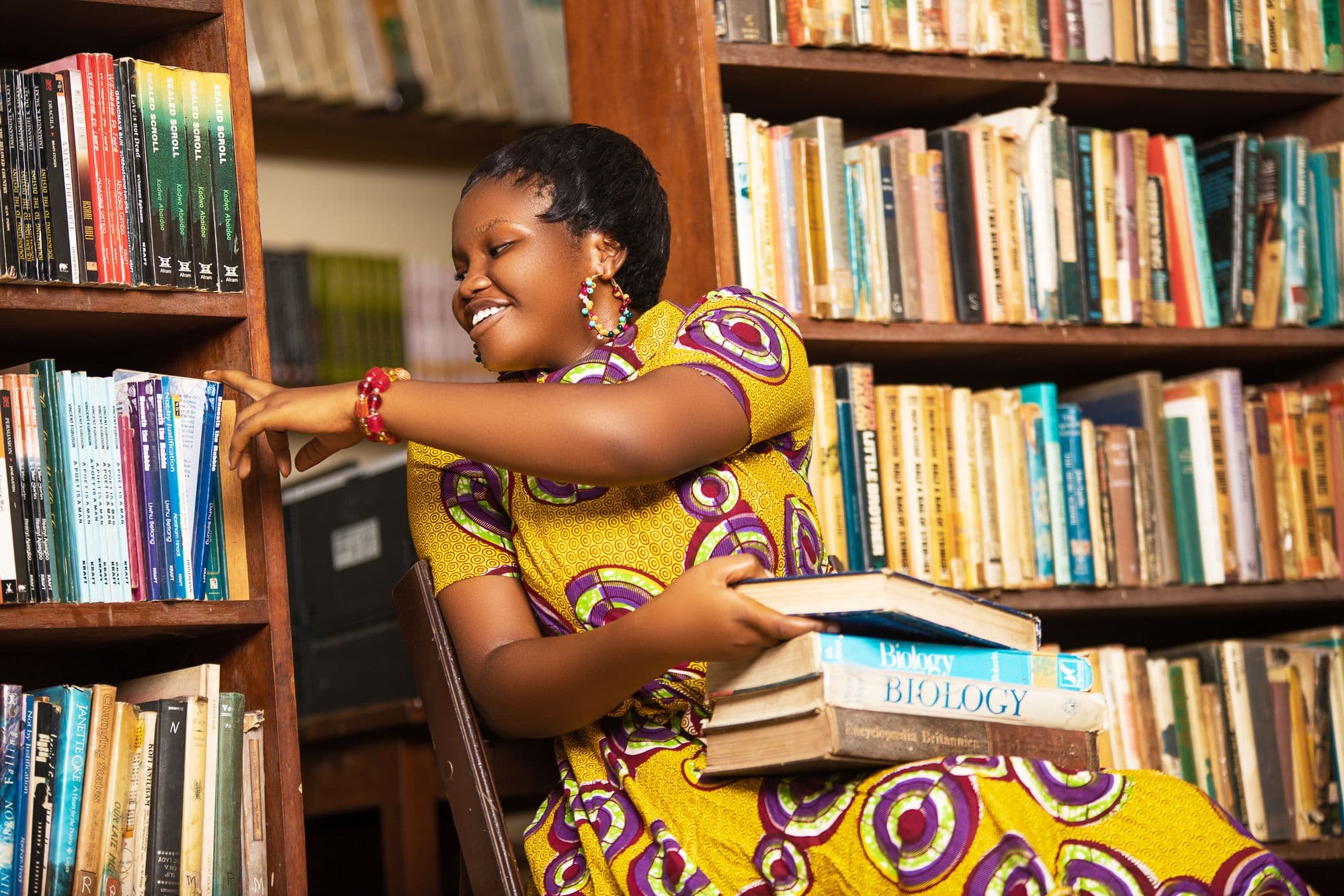 A woman in a yellow patterned dress choosing books from a library shelf in Ghana.